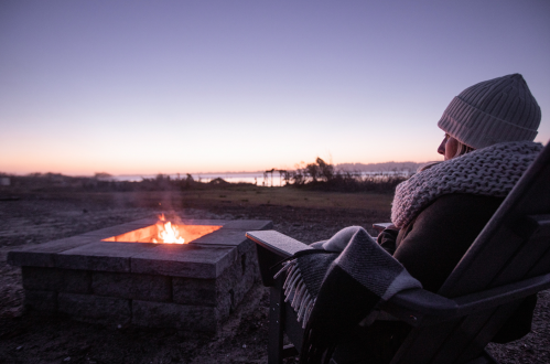 A person sits by a fire pit at dusk, wrapped in a blanket, with a serene landscape and water in the background.