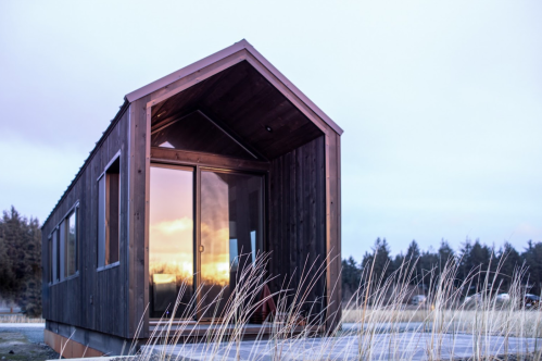A modern wooden cabin with a sloped roof, large glass doors, and a sunset reflection, surrounded by tall grass.