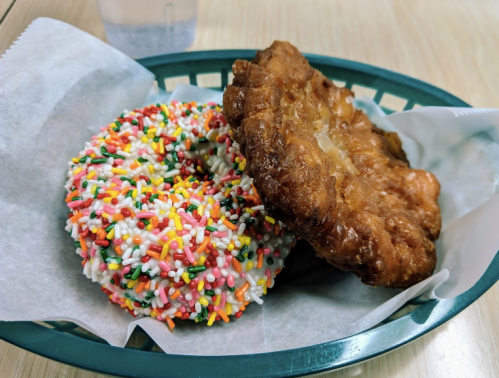A colorful sprinkle donut next to a golden-brown fried pastry in a green basket lined with white paper.