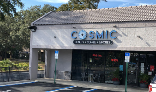 Exterior of Cosmic Donuts, Coffee & Savories shop with a drive-thru and open sign, surrounded by trees.