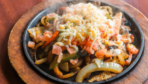 Sizzling fajitas on a hot plate, topped with melted cheese, tomatoes, and bell peppers, with steam rising.