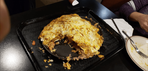 A large, cheesy dish with pulled meat and toppings, served on a black tray at a restaurant.