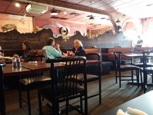 Two women sit at a table in a restaurant, engaged in conversation, with empty booths and a decorative wall in the background.