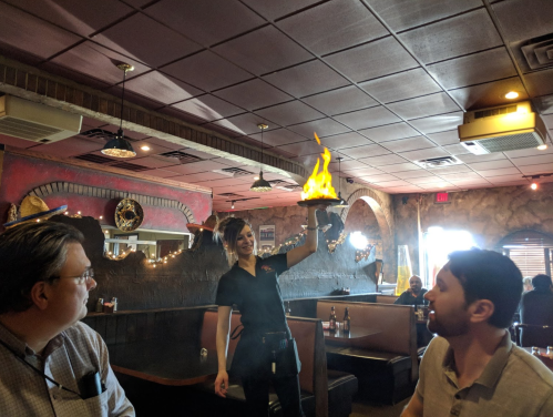 A waitress holds a flaming dish in a restaurant, while two diners watch in surprise.
