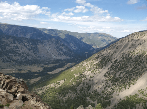 A panoramic view of mountains and valleys under a partly cloudy sky, showcasing lush greenery and rocky terrain.