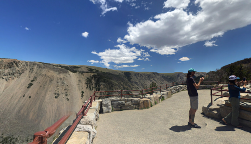Two people stand on a viewing platform overlooking a vast canyon under a blue sky with scattered clouds.