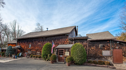 A rustic wooden building with a flag out front, surrounded by neatly trimmed bushes and a clear blue sky.