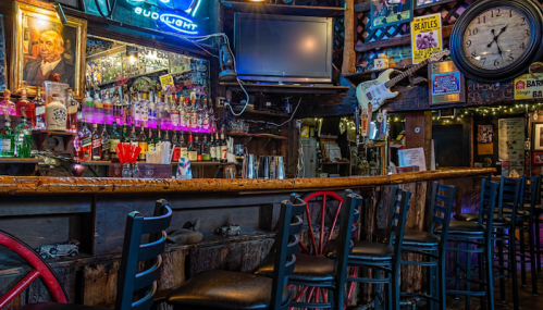 A cozy bar interior featuring a wooden counter, colorful liquor bottles, a neon sign, and vintage decor.