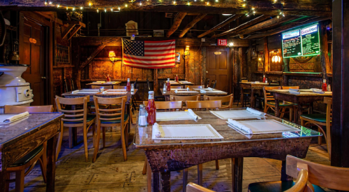 Cozy restaurant interior with wooden tables, chairs, an American flag, and a menu board illuminated by string lights.