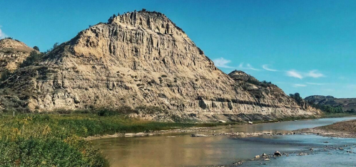 A rugged, sandy hill rises above a calm river, surrounded by green vegetation under a clear blue sky.