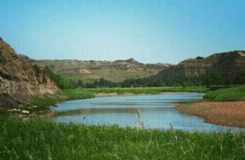 A serene river flows through lush green fields, surrounded by rocky hills under a clear blue sky.