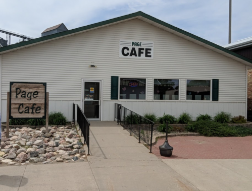 A white building with a green roof, labeled "Page Cafe," featuring a sign and landscaped entrance.