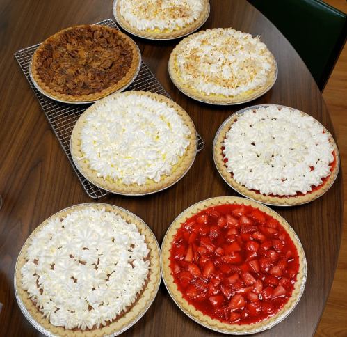 A table with eight delicious pies, including pecan, coconut cream, and strawberry, all topped with whipped cream.