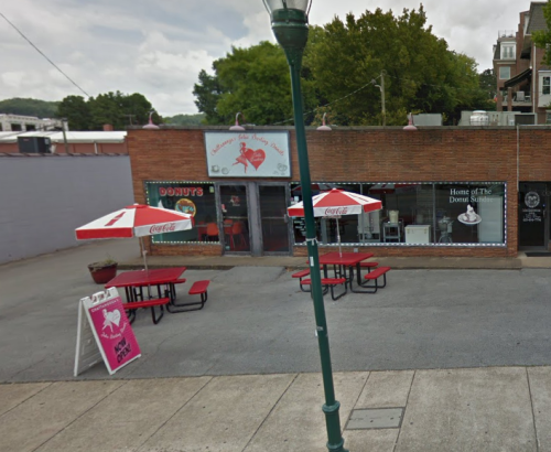 A donut shop with outdoor seating, featuring red tables and umbrellas, and a sign advertising "Home of the Donut Sundae."