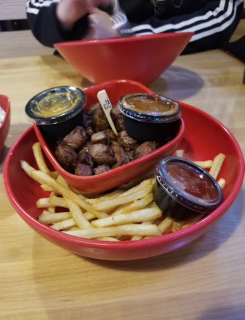A red bowl filled with fries and pieces of meat, accompanied by two small containers of sauce.