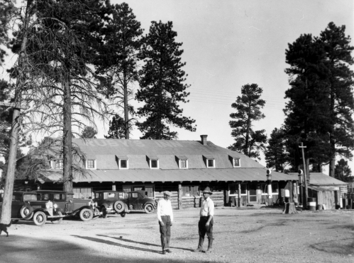 Two men walk in front of a rustic lodge surrounded by tall trees, with vintage cars parked nearby.