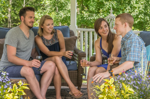 A group of four friends enjoying drinks and conversation on a porch surrounded by greenery and flowers.