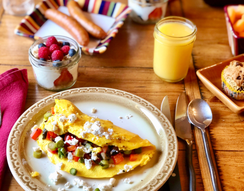 A colorful omelette with vegetables and feta cheese, served with fruit, yogurt, sausages, and juice on a wooden table.
