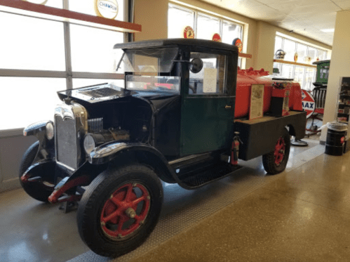 A vintage black and green truck with red wheels, parked indoors next to a gas pump and retro signage.