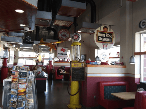A retro diner interior featuring vintage gas pumps, model airplanes, and colorful signage, with booths and a postcard display.