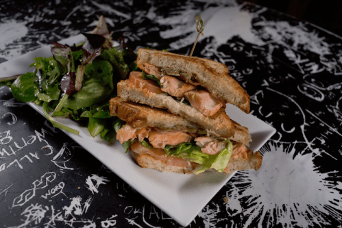 A stacked salmon sandwich with lettuce on toasted bread, served with a side of mixed greens on a patterned table.