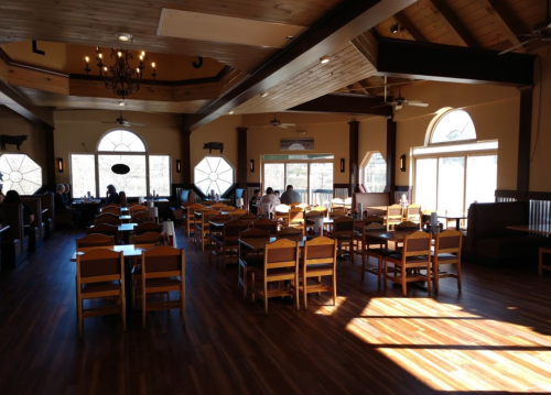 Interior of a spacious restaurant with wooden floors, large windows, and empty tables, illuminated by natural light.
