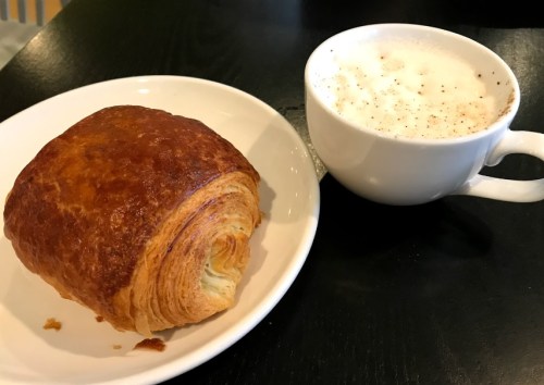 A golden-brown croissant on a white plate next to a cup of frothy cappuccino.