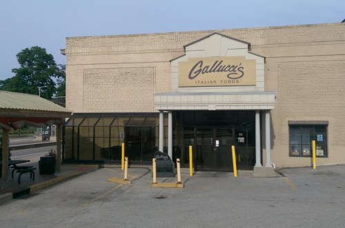 Exterior of Gallucci's Italian Foods, featuring a brick facade and large glass entrance, with parking and a bus stop nearby.