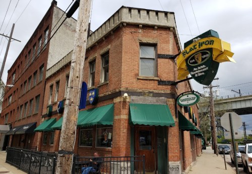 A brick building with green awnings, featuring a bar sign, located on a street with power lines and nearby structures.