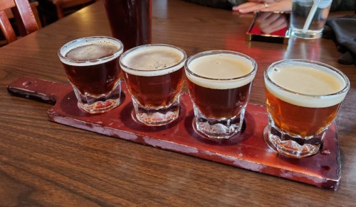 Four small glasses of beer on a wooden flight board, with a dark beer in the first glass and lighter beers following.