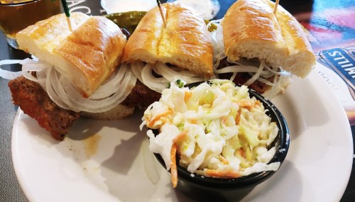 A plate with two sandwiches filled with fried meat and onions, accompanied by a small bowl of coleslaw.