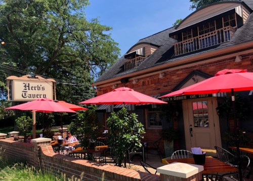 A brick building with a sign reading "Herb's Tavern," featuring red umbrellas over outdoor seating.