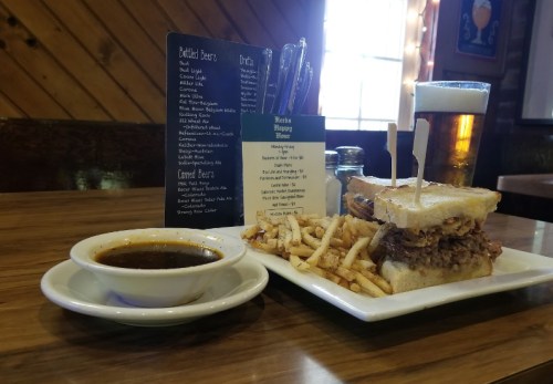 A plate with a sandwich, fries, and a bowl of soup, alongside a beer and a menu on a wooden table.