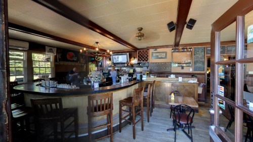 A cozy bar interior with wooden furniture, a curved counter, and shelves stocked with drinks. Natural light from windows.