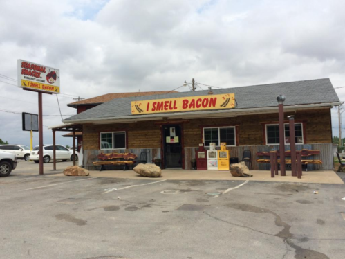 A rustic restaurant with a sign reading "I Smell Bacon," featuring outdoor seating and a gravel parking lot.