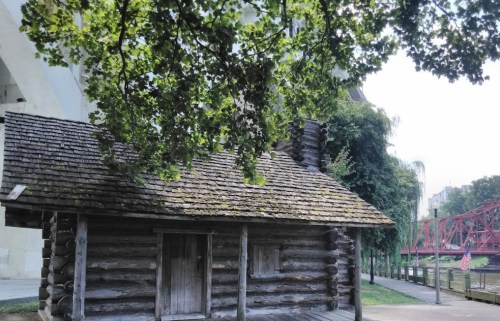 A rustic log cabin surrounded by trees, with a bridge and river in the background.
