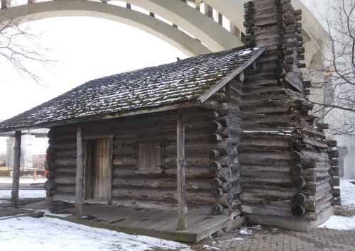 A rustic log cabin with a sloped roof, surrounded by snow, beneath a large concrete arch structure.