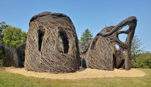 A large, abstract structure made of intertwined branches, set in a grassy area with trees in the background.