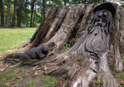 A carved wooden figure of a bearded man emerges from a tree stump, with a small animal sculpture nearby in a grassy area.
