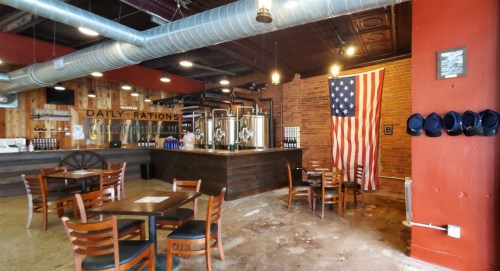 Interior of a rustic bar with brewing equipment, wooden tables, an American flag, and a warm, inviting atmosphere.