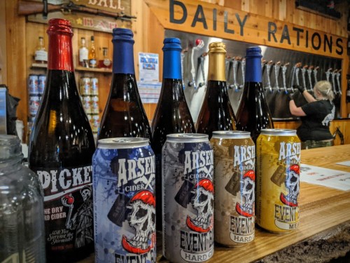 A variety of bottled and canned ciders displayed on a bar, with a bartender in the background.