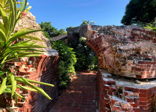 A pathway through weathered brick ruins, surrounded by lush greenery and blue sky.