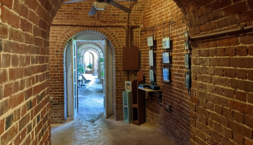 A brick hallway with arched doorways leading to a bright, open space with plants and artwork on the walls.