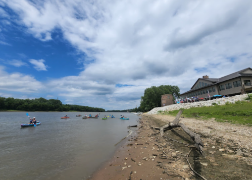 A river scene with kayakers paddling near a shore, featuring a building and trees under a partly cloudy sky.
