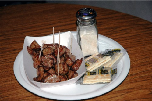 A plate of grilled meat cubes with toothpicks, accompanied by salt and crackers on a paper plate.