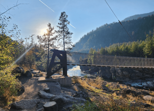 A suspension bridge spans a river, surrounded by trees and mountains, with sunlight filtering through the landscape.