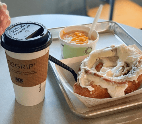 A coffee cup, a bowl of soup topped with cheese, and a large cinnamon roll with cream cheese frosting on a tray.
