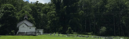 A small white church beside a cemetery, surrounded by lush green trees and a clear blue sky.
