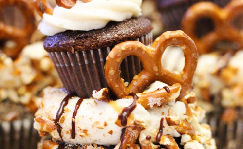 A close-up of a chocolate cupcake topped with frosting and a pretzel, surrounded by crushed pretzels and drizzled chocolate.