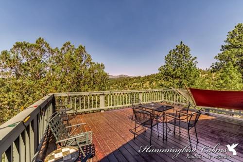 A scenic deck with chairs and a table, surrounded by trees and mountains under a clear blue sky.